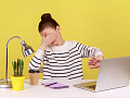 A young woman sitting at a desk in front of a yellow wall, puts one hand over her eyes and uses the other to shield her computer screen, suggesting 'I do not want to look at this'.